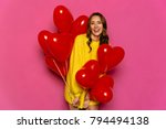 Charming young woman celebrating St. Valentine's day, holding red air balloons. Dressed in yellow sweater. Isolated over pink background.