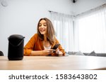 Small photo of Young cheerful woman controlling home devices with a voice commands, talking to a smart column at home. Concept of smart home and voice command control