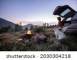 Woman enjoys beautiful view on the mountains, having a picnic with fireplace and sitting with dog in vehicle trunk at dusk on the evening. Traveling by car in nature