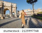 Small photo of Woman walks near Coliseum and Constantine Arch in Rome. Caucasian woman wearing summer dress and colorful shawl in hair. Concept of italian style and travel