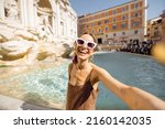 Cheerful woman taking selfie photo while visiting famous di Trevi fountain in Rome. Traveling Italy on a summer day concept. Portrait of caucasian woman in dress and shawl in hair