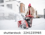 Woman removes snow with a snow thrower machine near house at residential area. Winter yard care and easy technology concept