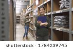 Small photo of female staff in warehouse office look to camera with clipboard in hands. smiling girl worker in rows of shelves in stockroom. coworker in back concentrated working and counting stocks with tablet.