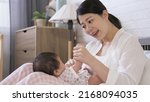 Small photo of asian first time mom is sitting and holding tiny hands with a smile while singing nursery song to her baby daughter on the bed in the bedchamber.