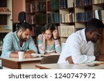 Small photo of Attentive multiracial male and female students sitting at a table in a public library while reading books and working over their coursework project.