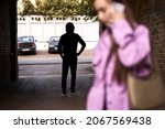 Criminal stalking a woman alone in dark street alley. Female is talking on phone. Unrecognizable man is hiding in the shadows. Woman Doesn