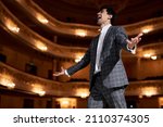professional opera singer in elegant classic outfit, singing hit with open mouth in microphone gesture with hand standing on stage in theater. Handsome caucasian guy during performance