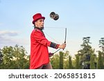 Side view of eccentric bearded man in red costume and hat juggling diabolo and looking at camera on blurred background of park