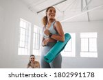 Smiling fitness woman standing in a fitness studio carrying a yoga mat. Portrait of a young woman at a fitness training centre.