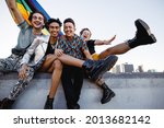 Small photo of Four LGBTQ people celebrating pride while sitting together. Four friends smiling cheerfully while raising the rainbow pride flag. Group of young queer individuals celebrating together outdoors.