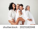 Female models of different ages celebrating their natural bodies in a studio. Four confident and happy women smiling cheerfully while wearing white shirts against a white background.