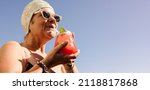 Small photo of Elderly woman drinking a tiki cocktail in swimwear. Carefree senior woman enjoying her summer vacation at a tropical spa resort. Happy mature woman enjoying herself after retirement.