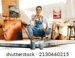 Small photo of Carefree female painter working in her art studio. Happy young artist smiling at the camera while squatting near her painting on the floor. Creative young woman making a new painting on a canvas.