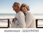 Small photo of Affectionate senior woman smiling happily while embracing her husband by the ocean. Romantic elderly couple enjoying spending some quality time together after retirement.