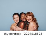 Three happy women with different skin tones smiling and embracing each other in a studio. Group of diverse women feeling comfortable in their natural skin. Body positive young women standing together.