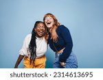 Small photo of Best friends laughing and having a good time together in a studio. Happy young women enjoying themselves while standing against a blue background. Two vibrant female friends making memories.