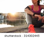 Young woman runner resting after workout session on sunny morning. Female fitness model sitting on street along pond in city. Female jogger taking a break from running workout.