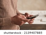 Small photo of Close-up of unrecognizable Black architect in sweater standing against table with sketch and using internet on smartphone