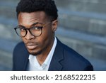 Small photo of Close-up of young Black businessman in eyeglasses raising eyebrow in puzzlement, he looking at camera outdoors