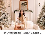 A pretty brunette young woman with a hairstyle in an elegant outfit holds a gift box in the decorated interior of the house during the Christmas holiday and New Year indoor. Selective focus
