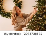Small photo of A joyful smiling young woman is preparing for the Christmas holiday, decorating a Christmas tree and holding gift boxes in a cozy interior room of the house. Selective focus