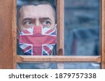 Small photo of Young man in a protective mask with a British flag stays at home looking out the window. Lockdown in the UK. Corona virus mutated covid-19. Strengthening quarantine. Coronavirus is mutating. Health