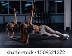 Small photo of Two beautiful women with athletic figures perform a side bar in a dark gym. Two female athletes make balance from yoga.