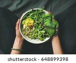 Green vegan breakfast meal in bowl with spinach, arugula, avocado, seeds and sprouts. Girl in leggins holding plate with hands visible, top view. Clean eating, dieting, vegan food concept