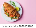 Croissant sandwich with cream cheese, salmon and arugula on a white plate, pink background, copy space, top view. Healthy breakfast concept.