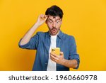 Amazed shocked caucasian guy holding smartphone in his hand, looking at the phone in surprise with his glasses raised, stunned facial expression, stands on isolated orange background