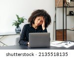 Upset depressed young adult african american woman, top manager, office worker, director of the company, sits at the desktop in front of the laptop, looks at the screen in disappointment, got bad news