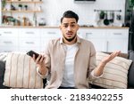 Confused puzzled indian or arabian guy in casual clothes, sits on a sofa in an interior living room, holds a smartphone in his hand, looks questioningly at the camera, spreading his arms around