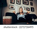 Small photo of Woman Sitting on a Sofa Covering her Ears with Pillows. Unhappy lady suffering from a headache due to loud neighbors and thin walls