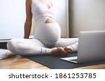 Closeup belly of pregnant woman practicing yoga online at home with laptop. Expectant mother doing prenatal yoga class or meditating during pregnancy indoors. Healthcare of mother and unborn baby.