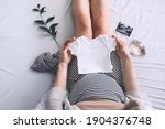 Pregnant woman holding white baby bodysuit preparing to child birth during pregnancy. Young mother with pregnant belly waiting of a baby.