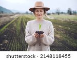Young plant in hands in background of agricultural field area. Woman holding in hands green sprout seedling on black soil. Concept of Earth day, organic gardening, ecology, sustainable life.