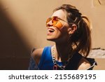 Small photo of Attractive woman with high ponytail standing near beige wall in old city. Girl joy of sun light, wear orange sunglasses look at sun. Shadow and light of sunny summer. Focus on shackle of sunglasses.