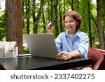 Adorable mature woman with laptop sitting at the table in outdoor cafe in the park. Middle-aged smiling brunette in a blue shirt works and plays using modern technology. Active lifestyle for all ages.