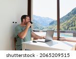 Man working from his apartment in the mountains. He is drinking coffee while is looking through the window during a work break. Working remotely.