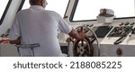Small photo of The captain of a pleasure boat sits at the helm and follows the course of the ship. The right hand holds the helm, and the captain looks into the distance. White cabin of the captain.