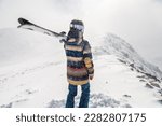The skier stands on the top of the mountain and enjoys the view of the beautiful winter mountains on a cloudy day. Woman in ski suit holding skis on her shoulder