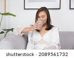 Small photo of Asian woman in white nightgown drinking warm water on the bed at living room.