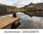 Young woman wearing a woolly hat sitting on the edge of a pier overlooking the mirror-like water of the Loch Leven with mountains on the background and boats floating on the water in Glen Coe