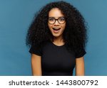 Small photo of Pleasantly surprised dark skinned woman keeps jaw dropped, gazes with interest, has curly hair, wears black t shirt, models against blue wall. Afro American lady gazes with shock, has bated breath
