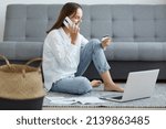 Full length portrait of happy exited woman wearing white shirt and jeans sitting on floor near sofa, using laptop and talking phone, holding credit card and expressing happiness.