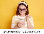 Small photo of Horizontal shot of confused young woman wearing striped t-shirt standing with mobile phone and looking at display with puzzlement, posing isolated over yellow background.