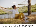 Outdoor shot of Caucasian young adult woman sitting on hammock by the water, looking away, resting, spreads hands, laughing happily, wearing white shirt and yellow trousers.