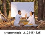 Small photo of Outdoor shot of young positive couple sitting in the forest with overhead projector with empty white screen with advertisement area, family holding hands or giving high five.