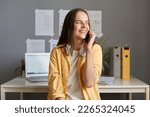 Small photo of Photo of dreaming smiling woman with brown hair, fem ale wearing casual clothes and has headphones, talking via cell phone and looking away with happy expression.