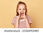 Excited little brown haired girl wearing striped t-shirt standing isolated over beige background keeps hands near mouth screaming loud important information.